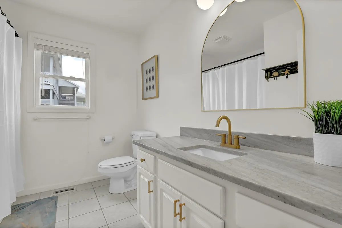 A calming bathroom scene featuring a vanity with a sink, a large mirror, soft natural light from a window, framed artwork, and a lush plant, creating a welcoming space for personal care routines.