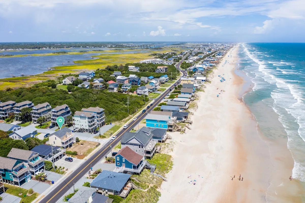 A wide-angle exterior view featuring beachfront properties, the ocean stretching out to the horizon, a road leading directly to the beach, and lush surrounding greenery. Several groups can be seen enjoying the sand and surf, suggesting a lively atmosphere ideal for water sports and group activities.
