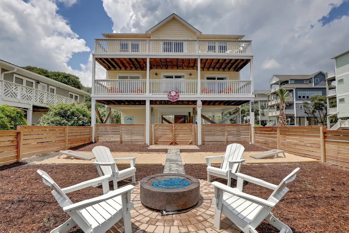 A three-story beach house stands behind a securely fenced yard with a sandy play area. Adirondack chairs surround a fire pit, and palm trees add a tropical touch. The fence is clearly visible, creating a safe, enclosed space ideal for both children and pets. The overall vibe is relaxed and sociable, with space for outdoor activities and family gatherings.