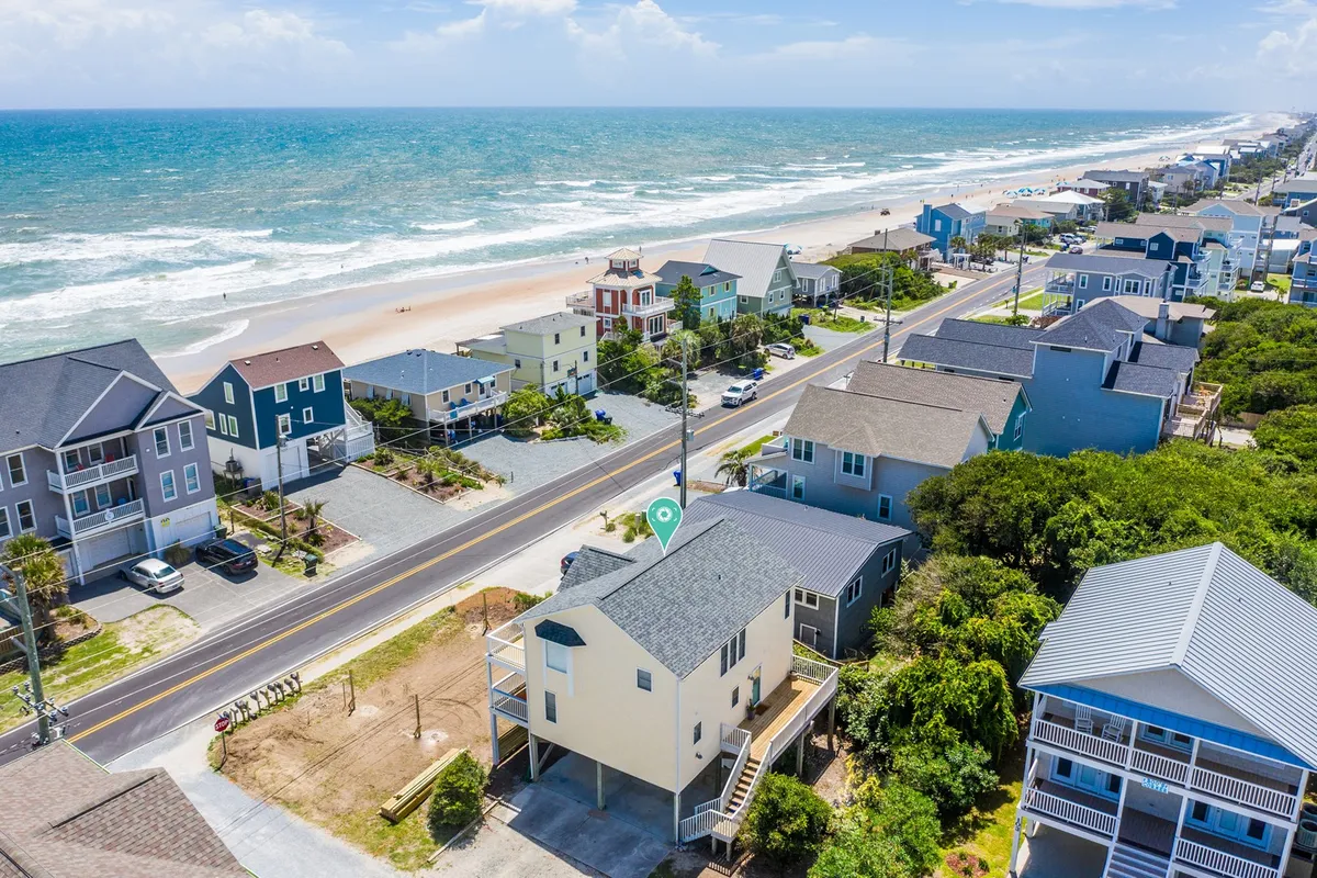 A panoramic exterior shot of the beachfront with the ocean in the foreground and houses lining the coastline. The scene is relaxed, with visible activity on the sand, evoking the spirit of outdoor adventure and group enjoyment.