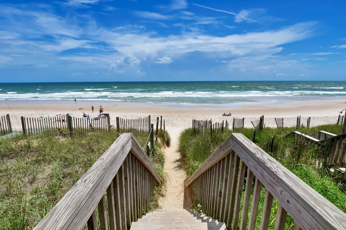 A lively scene showing wooden stairs leading directly to the beach, with people enjoying the sand and surf in the background. Dune grass adds a natural touch, and the setting feels joyful and family-friendly.