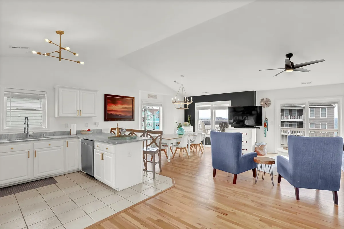 A spacious, light-filled interior featuring a kitchen island, dining area, blue armchairs, and large windows looking out onto the natural surroundings. The open layout is ideal for cooking, dining, and relaxing with friends after a day of adventure.