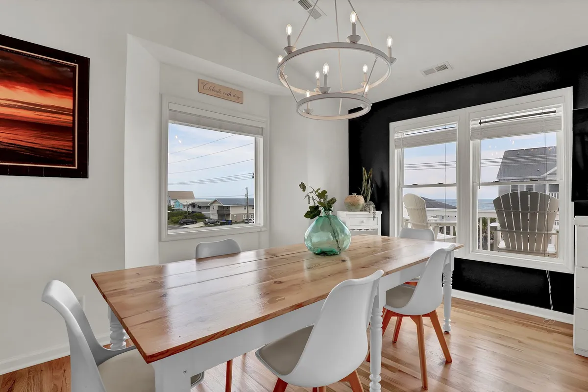 A welcoming dining area is highlighted here, with a large table beneath an elegant chandelier, surrounded by windows that offer glimpses of the outdoors. The room's layout encourages shared seaside meals, games, and memorable gatherings, capturing the friendly, communal vibe promised in the text.