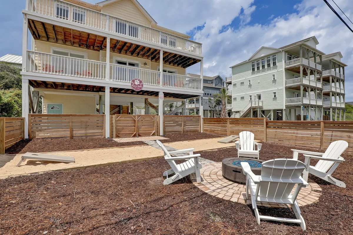 A two-story house with balconies overlooks a fenced yard, where white Adirondack chairs encircle a fire pit. The yard features a sand area, and the fence ensures a safe environment for both pets and children. The scene is lively and social, evoking a sense of community and fun.