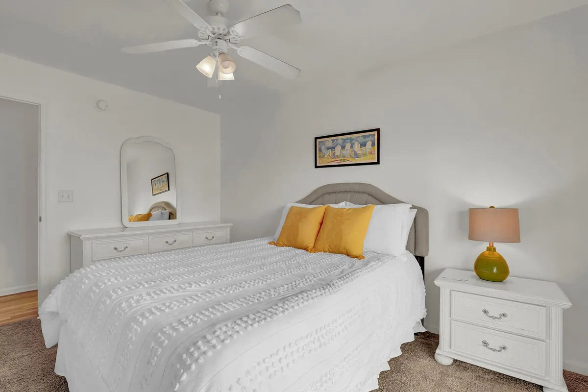 A peaceful bedroom with white bedding accented by yellow pillows, a nightstand with lamp, a mirror, and framed artwork. The mood is cozy and inviting, suggesting a restful space to unwind.