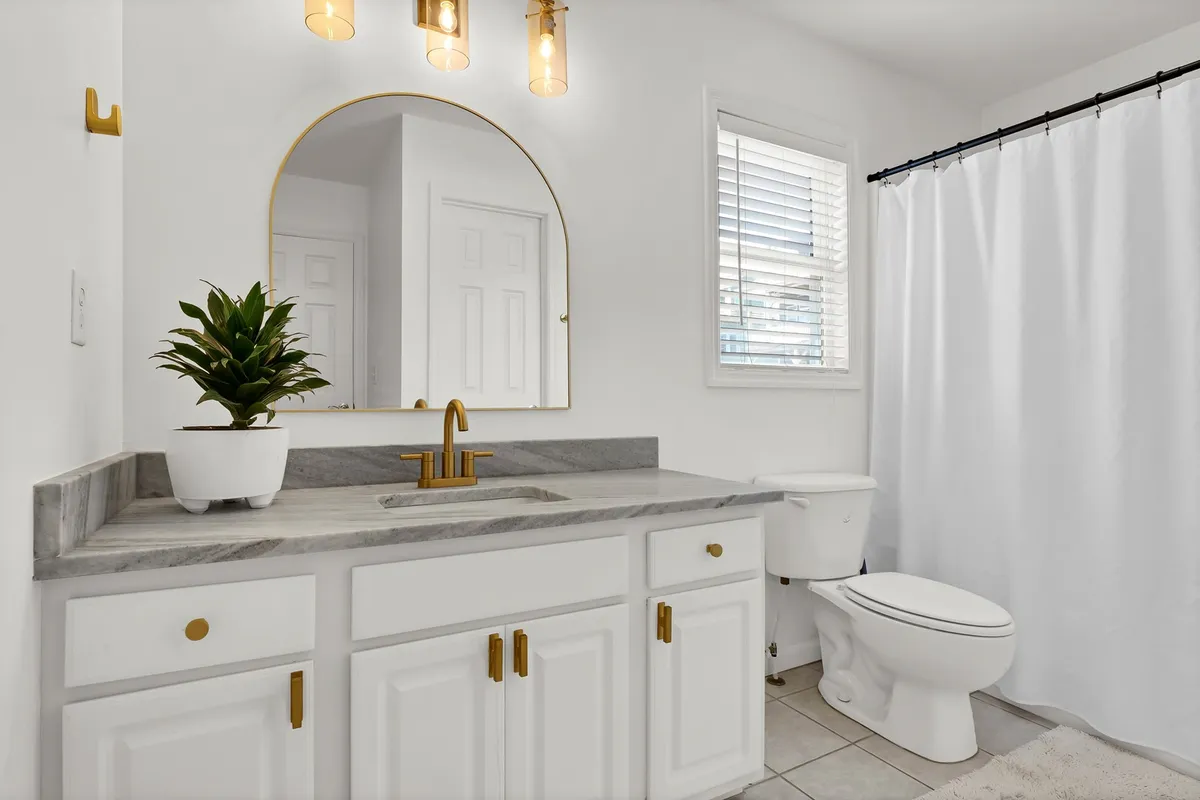 A modern, serene bathroom featuring elegant gold fixtures, a large mirror above a sleek vanity, a touch of greenery, and a pristine white shower curtain. The space feels calm and designed for self-care.