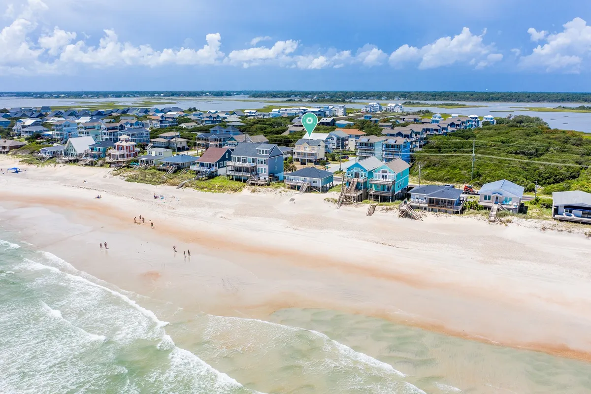 A wide exterior beach scene featuring beachfront houses, a sandy beach, gentle ocean waves, and people walking along the shore under a cloudy sky. The image conveys a leisurely and carefree mood, highlighting the accessibility of both the beach and local attractions.
