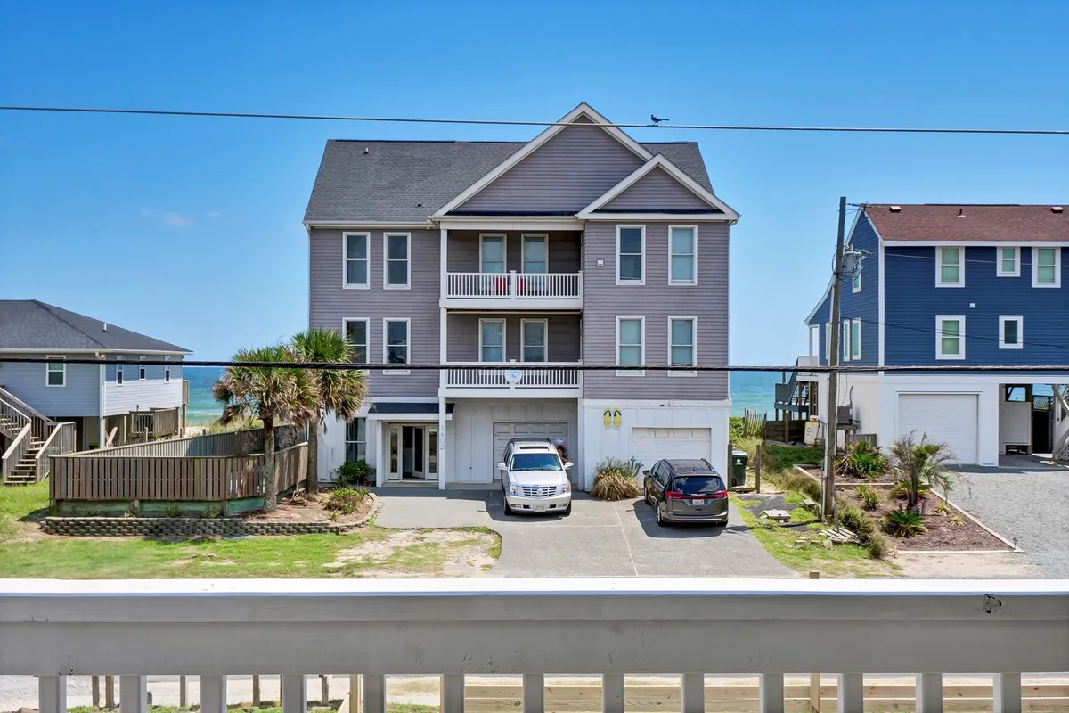 An exterior view of a beachfront property with balconies, cars in the driveway, and the beach visible nearby. The scene feels relaxed and welcoming, suggesting ease of parking and immediate beach access for spontaneous adventures.