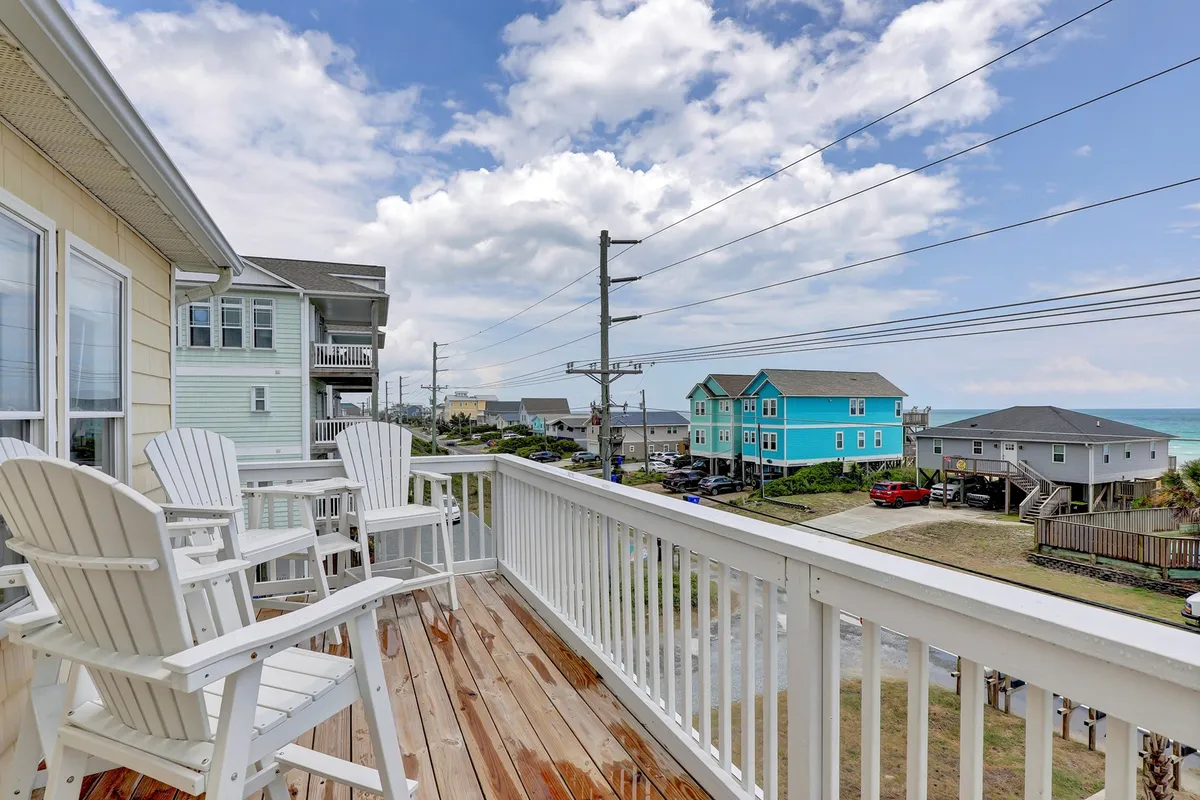A cheerful exterior scene showing balcony seating with views of the ocean and colorful houses. The setting is casual and inviting, with space for friends to relax while overlooking the lively beach neighborhood, reflecting the energy and convenience of the downtown Surf City area.