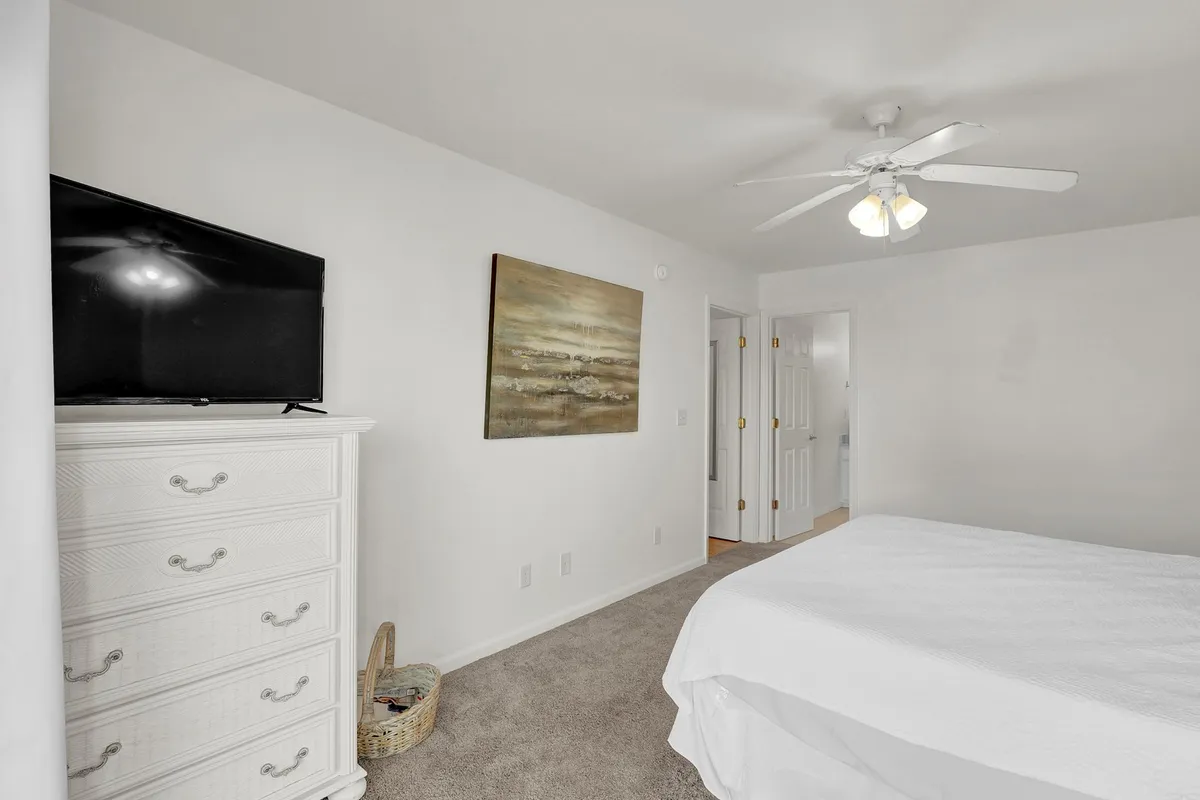 A serene bedroom featuring a bed with crisp white linens, a white dresser, a flat-screen TV, and an abstract painting. The room is filled with natural light and has a peaceful, uncluttered ambiance.