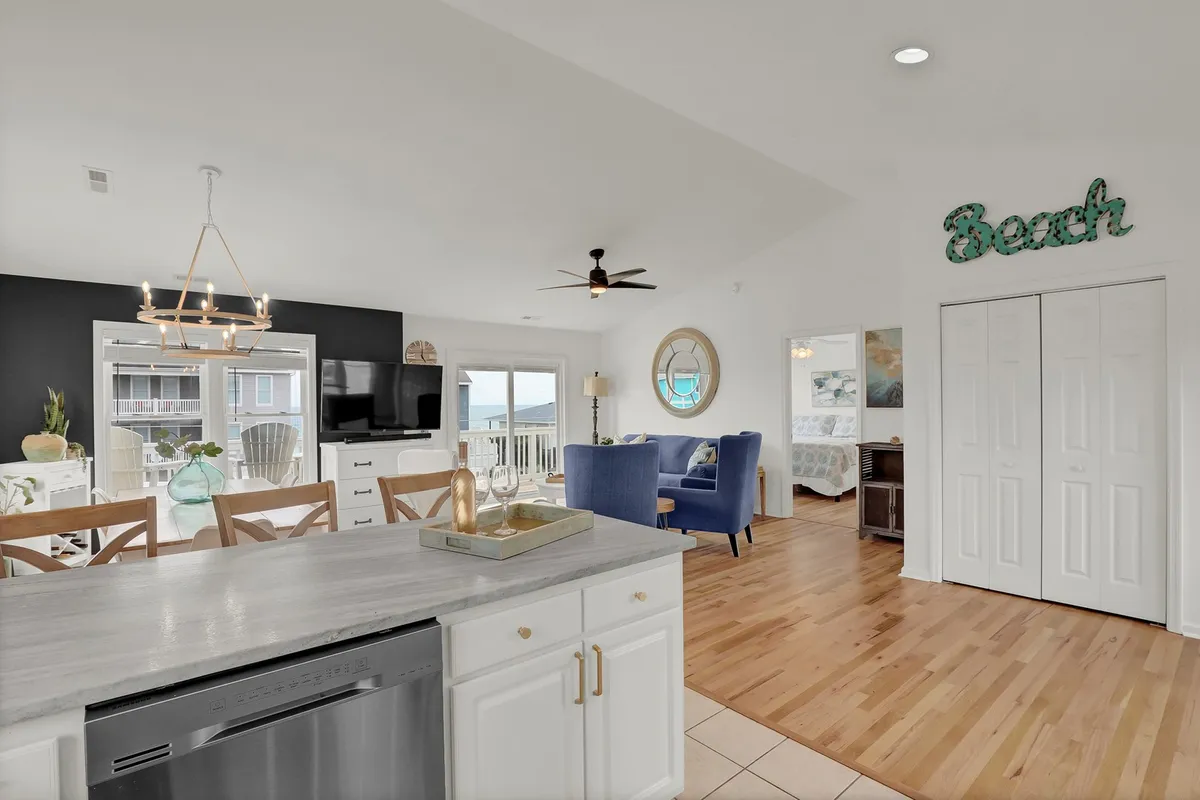 A modern kitchen island with bright decor and playful blue accent chairs is the focal point here. The design encourages casual dining and socializing, with easy access to both the kitchen and adjoining living spaces. The bright 'Beach' sign and decor add a cheerful, coastal touch.