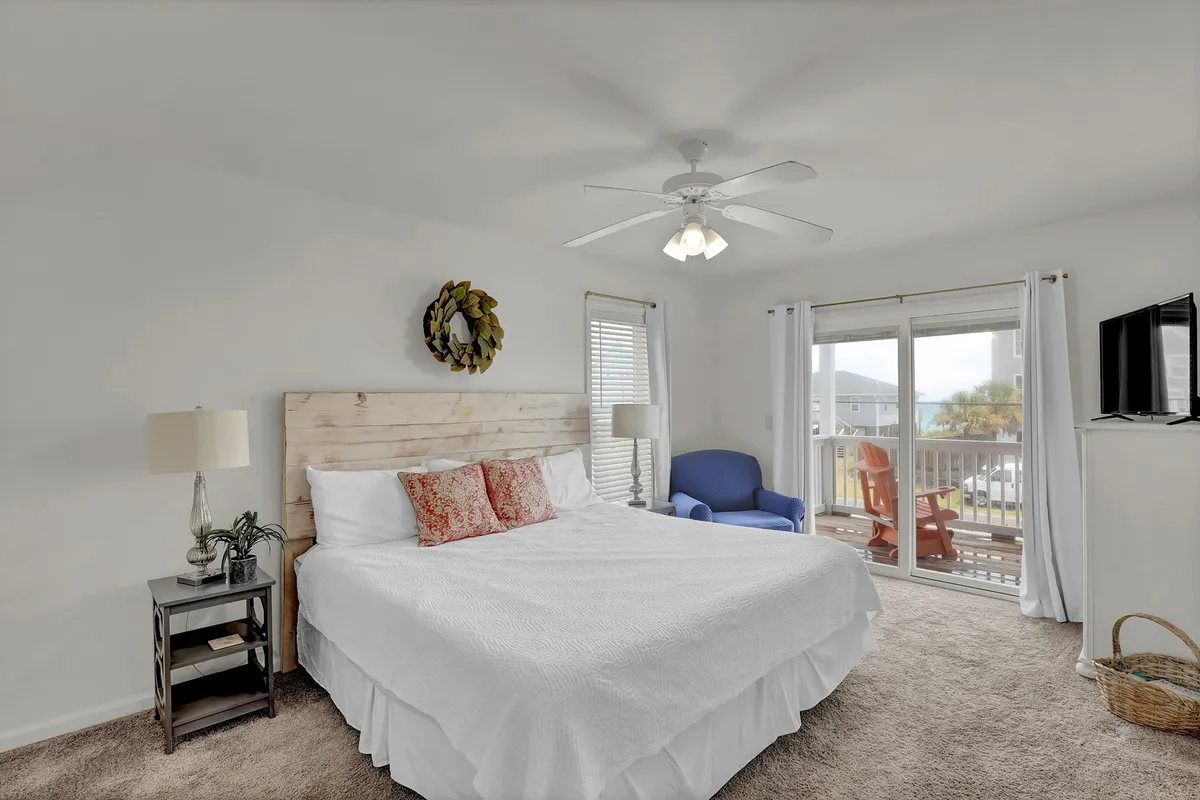 A bedroom with a plush bed dressed in crisp white linens sits next to a blue accent chair and a wooden headboard. A decorative wreath adds a touch of coastal charm above the bed, and a television is mounted for cozy evenings. Soft natural light filters in, creating a tranquil ambiance.